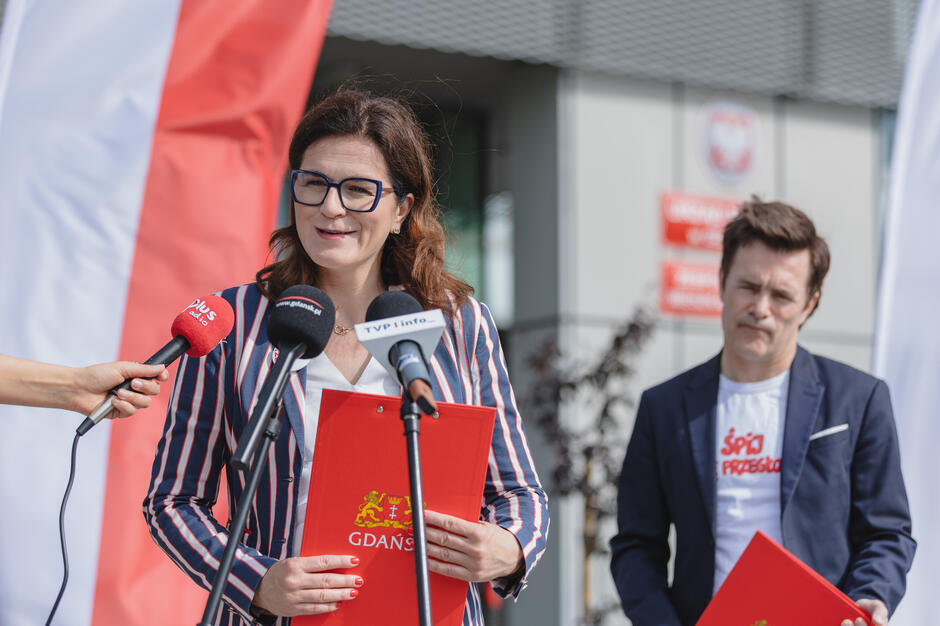 Kobieta w marynarce w paski przemawia na konferencji prasowej w Gdańsku, trzymając czerwoną teczkę z herbem miasta, a obok niej stoi mężczyzna z podobną teczką. 