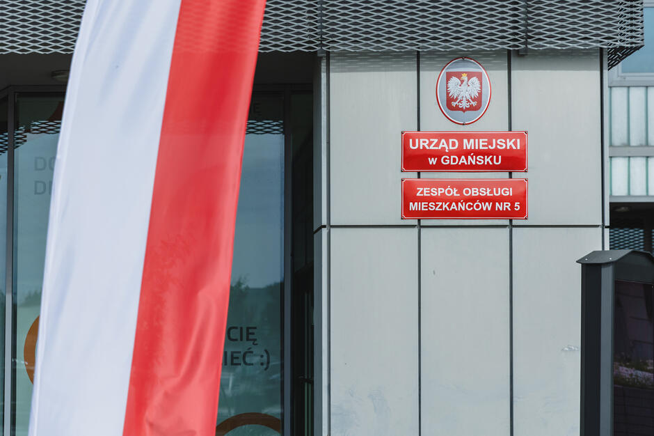 =Na zdjęciu widoczna jest fasada Urzędu Miejskiego w Gdańsku – Zespół Obsługi Mieszkańców nr 5, obok której powiewa flaga Polski. 