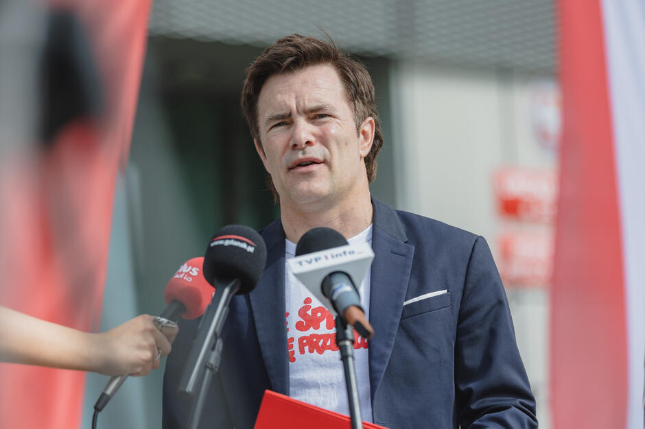  mężczyzna stoi za podium z wieloma mikrofonami przed sobą, nosi niebieski blezer i białą koszulkę z czerwonym logo na sobie, człowiek wydaje się mówić do jednej z osób widocznych po lewej stronie jego wizerunku, trzymając mikrofon i przemawiając do mediów, w tle czerwono-białe transparenty z logo firmy lub organizacji, wydaje mi się, że człowiek wypowiada przemówienie lub zwraca się do publiczności.