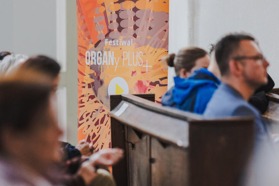 Fotografia przedstawia uczestników wydarzenia siedzących w drewnianych ławkach, na tle kolorowego baneru z napisem „Festiwal ORGANy PLUS+”. Atmosfera sugeruje skupienie i oczekiwanie przed rozpoczęciem koncertu.
