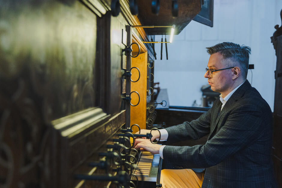 Zdjęcie ukazuje organistę w eleganckim garniturze grającego na klasycznych organach, z pełnym skupieniem na instrumencie. Oświetlenie podkreśla detale zabytkowego stołu gry oraz koncentrację muzyka.