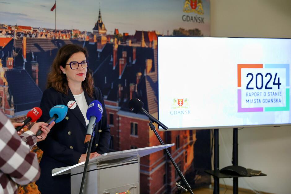 na zdjęciu kobieta w średnim wieku, mówi do dziennikarskich mikrofonów