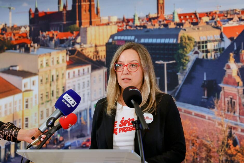 na zdjęciu kobieta w średnim wieku, z białą koszulką i czerwonym napisem, mówi do mikrofonów