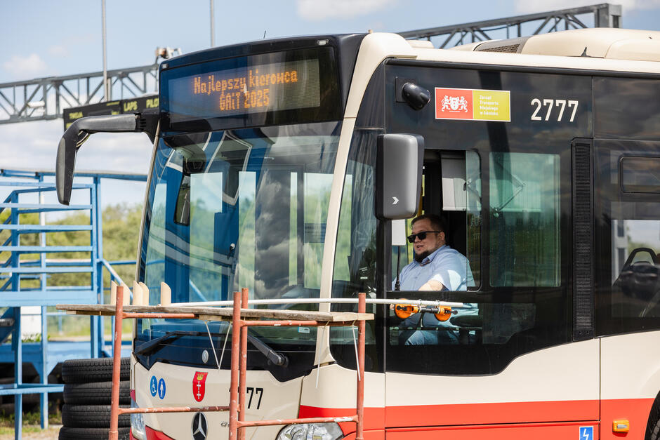Na zdjęciu widzimy kierowcę miejskiego autobusu biorącego udział w konkursie „Najlepszy kierowca GAiT 2025”, co potwierdza napis na wyświetlaczu pojazdu. Uśmiechnięty mężczyzna w okularach przeciwsłonecznych siedzi za kierownicą autobusu, gotowy do jazdy po specjalnym torze. 