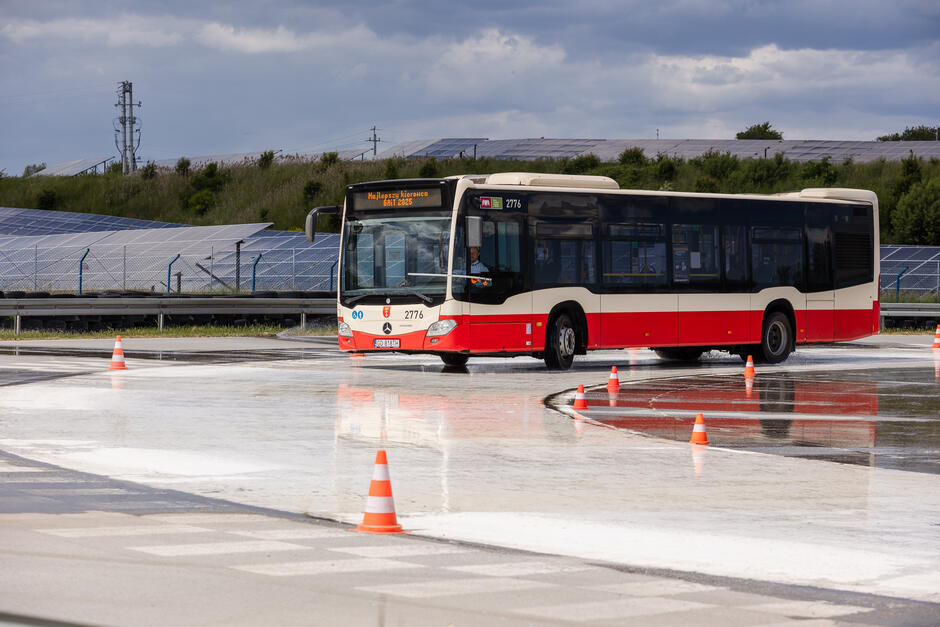 Na zdjęciu widzimy autobus komunikacji miejskiej jadący po mokrym torze manewrowym, oznaczonym pomarańczowymi pachołkami. Na wyświetlaczu autobusu widnieje napis „Najlepszy kierowca GAiT 2025”, co sugeruje, że pojazd bierze udział w konkursie sprawnościowym. 