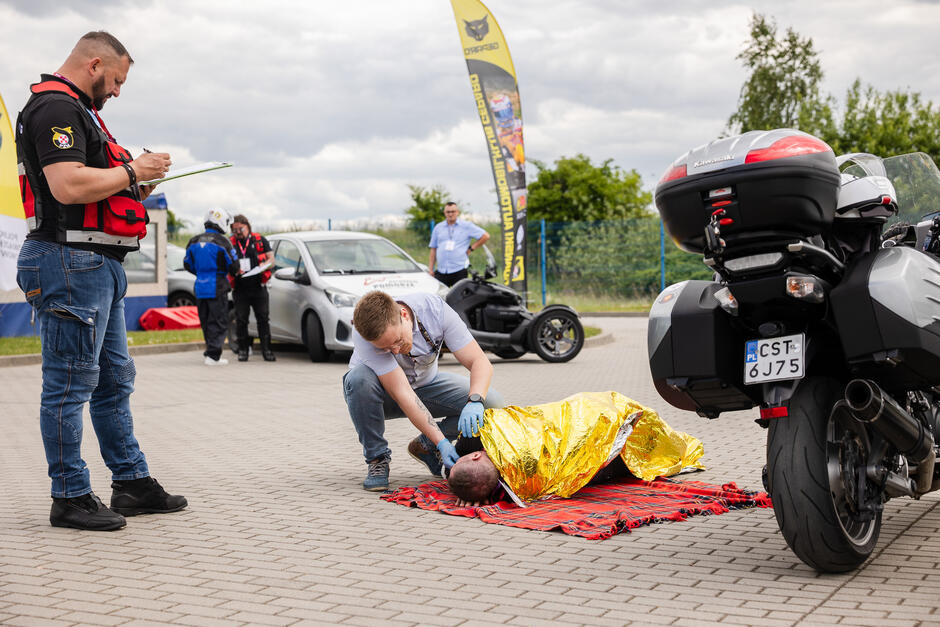 Na zdjęciu przedstawiono symulację akcji ratunkowej po wypadku drogowym – ratownik udziela pomocy poszkodowanemu owiniętemu w koc termiczny. W tle widoczni są organizatorzy i obserwatorzy wydarzenia, a także pojazdy, w tym motocykl i samochód oznakowany jako szkoła jazdy.
