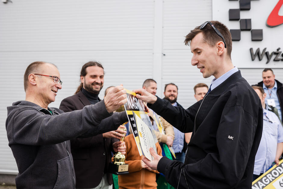 Na zdjęciu uwieczniono moment wręczania nagrody jednemu z uczestników konkursu, który z uśmiechem odbiera dyplom i symboliczny czek. W tle zgromadzeni ludzie gratulują zwycięzcy, a jeden z nich trzyma puchar, co podkreśla uroczysty charakter wydarzenia.
