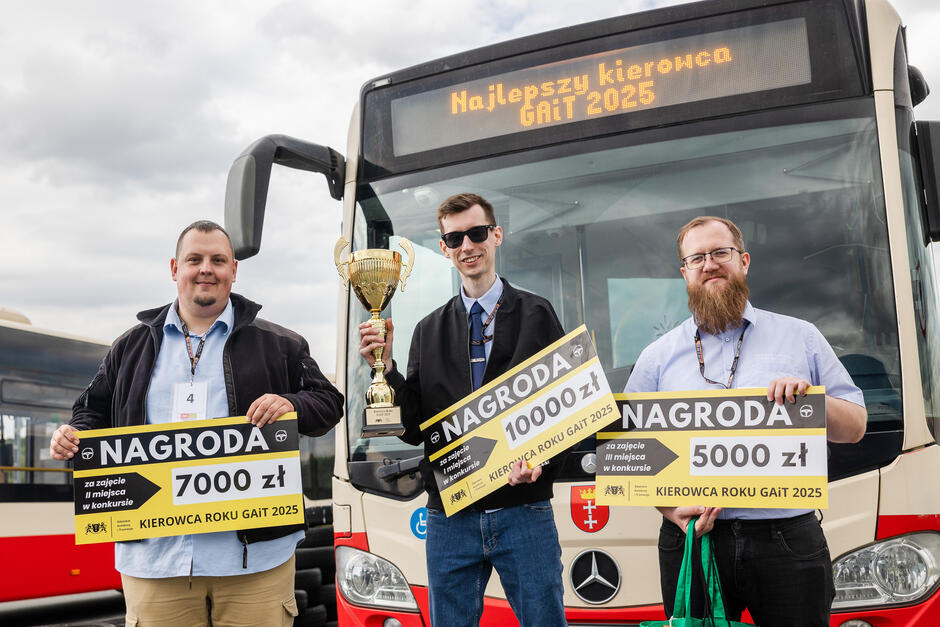 Na zdjęciu trzej zwycięzcy konkursu „Kierowca Roku GAiT 2025” z dumą prezentują swoje nagrody pieniężne oraz puchar, stojąc przed miejskim autobusem z napisem „Najlepszy kierowca GAiT 2025”. Uśmiechnięci i elegancko ubrani laureaci trzymają tablice z informacją o zajętych miejscach i wysokości wygranych: 10 000 zł, 7 000 zł oraz 5 000 zł. 