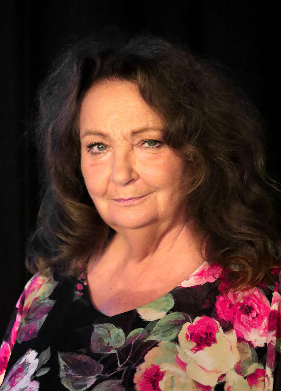 Zbliżenie tej samej kobiety ukazuje ją z bardziej wyrazistym wyrazem twarzy, z lekkim przymrużeniem oczu, co nadaje jej spojrzeniu pewności siebie i doświadczenia. Oświetlenie podkreśla rysy twarzy, a kolorowa, kwiecista bluzka pozostaje tym samym wyrazistym elementem garderoby