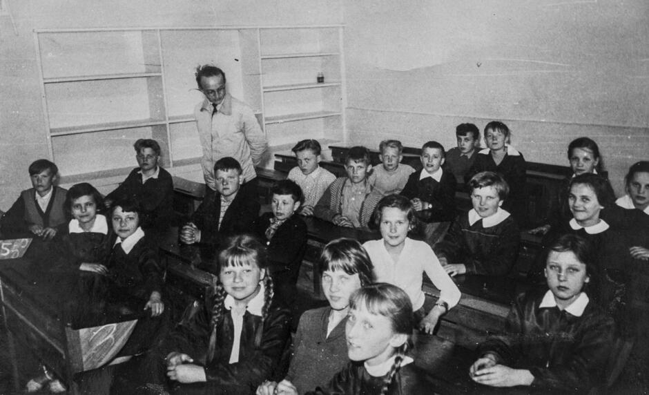 To czarno-białe zdjęcie przedstawia scenę z sali lekcyjnej z przeszłości, prawdopodobnie z lat 50. lub 60. XX wieku w Polsce. W klasie znajduje się grupa uczniów – zarówno chłopców, jak i dziewcząt – siedzących w drewnianych ławkach. Większość uczniów ma na sobie szkolne fartuszki z białymi kołnierzykami, co było typowe dla ówczesnego stroju uczniowskiego.