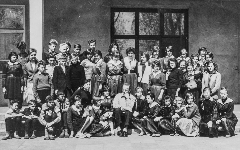 To czarno-białe zdjęcie grupowe przedstawia dużą grupę uczniów wraz z nauczycielem, wykonane zapewne na dziedzińcu szkolnym, przed wejściem do budynku. Scena pochodzi najprawdopodobniej z lat 50. lub 60. XX wieku.