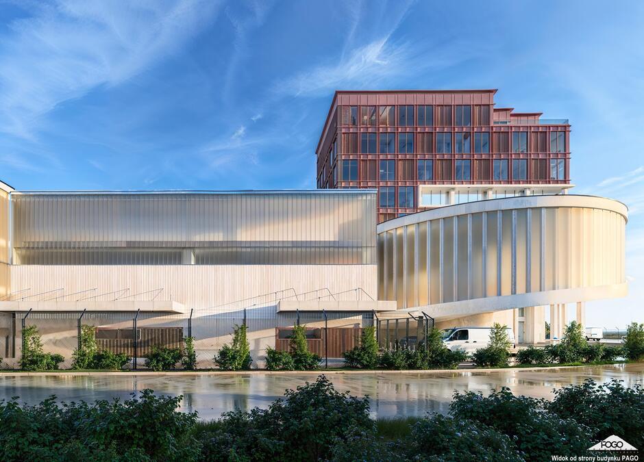 Na zdjęciu widoczny jest nowoczesny budynek o zróżnicowanej bryle architektonicznej, łączący przeszklone i metaliczne elementy z geometrycznymi formami. W dolnej części znajduje się rampa wjazdowa i zaparkowany biały samochód dostawczy, a całość otacza ogrodzenie z zielenią, co sugeruje funkcję użytkowo-administracyjną, prawdopodobnie punkt kontroli granicznej.