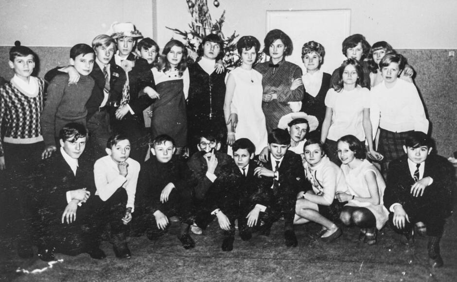 To czarno-białe zdjęcie przedstawia grupę młodzieży szkolnej, pozującej wspólnie do pamiątkowej fotografii we wnętrzu szkoły. W tle widoczna jest choinka, co sugeruje, że zdjęcie zostało wykonane podczas zabawy świątecznej lub noworocznej – być może klasowego spotkania opłatkowego lub choinki szkolnej. Uczniowie są ubrani odświętnie – chłopcy w garniturach lub swetrach, a dziewczęta w sukienkach, spódnicach i eleganckich bluzkach.