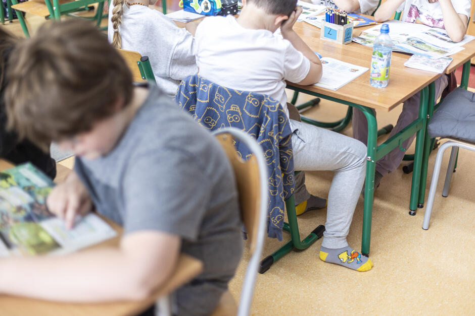 Zdjęcie przedstawia wnętrze klasy szkolnej, w której dzieci siedzą przy ławkach i pracują z podręcznikami. Uczniowie są pochłonięci nauką – jeden z nich intensywnie czyta, inny podpiera głowę na ręce, a obok na biurkach widoczne są otwarte książki, zeszyty, przybory i butelki z napojami. Uwagę przyciągają detale – kolorowe skarpetki z motywem konika oraz bluza przewieszona przez oparcie krzesła z nadrukiem postaci w stylu kreskówkowym. W tle widać klasyczne zielone krzesła i stoły z drewnianymi blatami, typowe dla polskich szkół podstawowych.