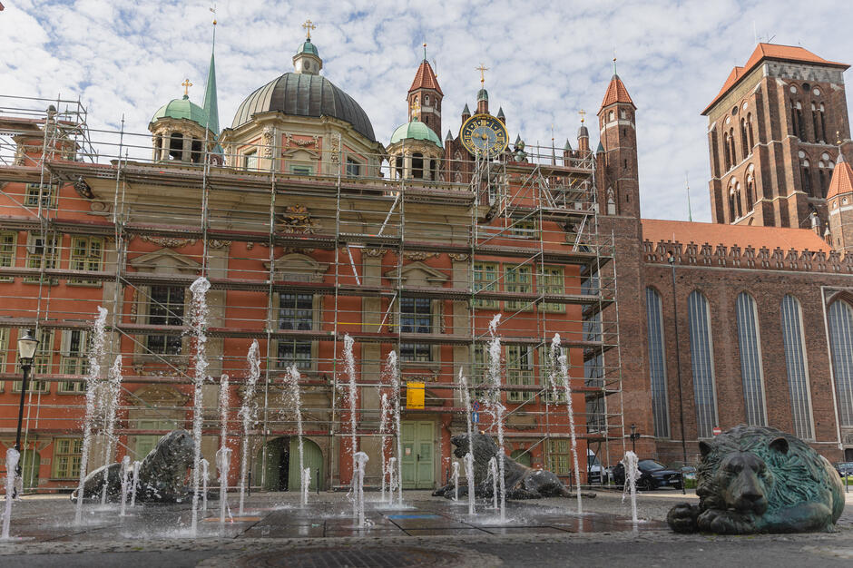 Na pierwszym planie widoczna jest fontanna z rzeźbami lwów, a za nią kaplica ozdobiona rusztowaniami, co sugeruje trwające prace konserwatorskie. W tle wznoszą się wieże Bazyliki Mariackiej w Gdańsku.