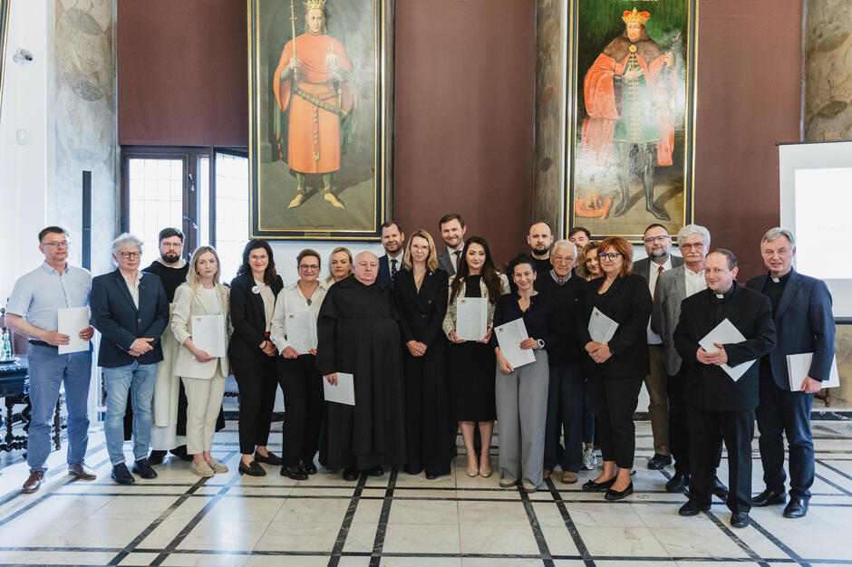 Grupa osób pozuje do wspólnego zdjęcia w reprezentacyjnej sali z dużymi portretami historycznych postaci, trzymając dokumenty — prawdopodobnie akt przekazania dotacji lub wyróżnienia. Wnętrze ma charakter historyczny i uroczysty.