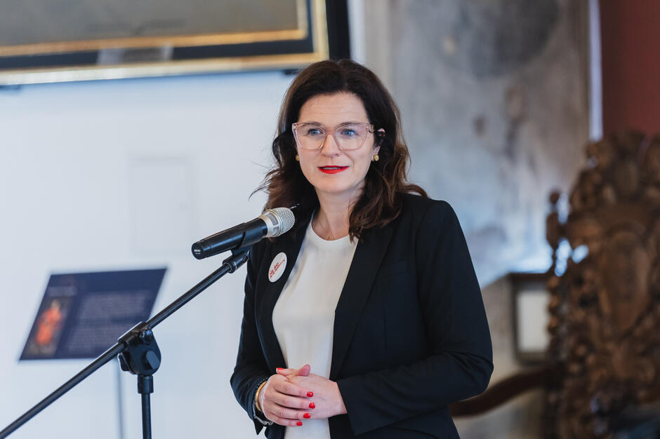 Kobieta w ciemnym garniturze przemawia do mikrofonu, stojąc w eleganckim, zabytkowym wnętrzu z dekoracyjnym krzesłem w tle. Ma przypinkę z napisem „Gdańsk”, co sugeruje, że reprezentuje lokalne władze.


