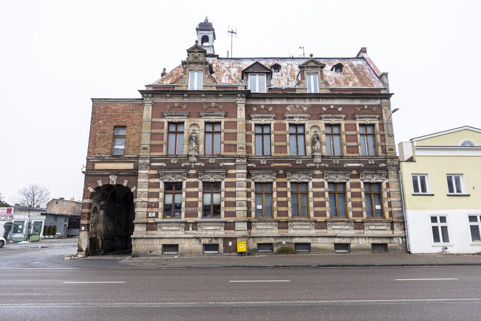 Stara, bogato zdobiona kamienica z czerwonej cegły i kamienia stoi przy głównej ulicy, z widocznymi oznakami zużycia i uszkodzeń. Obok niej znajduje się współczesny budynek mieszkalny, co uwypukla kontrast między historią a nowoczesnością.