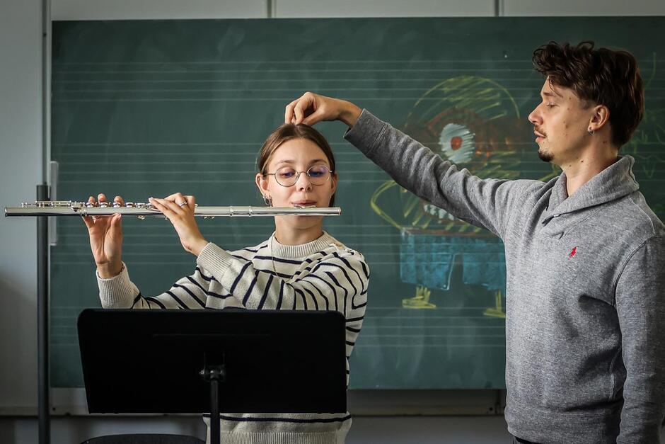 Pośrodku zdjęcia stoi dziewczynka grająca na flecie, po lewej jest nauczyciel, który trzyma dłoń na czubku jej głowy. W tle jest zielona tablica z narysowanym krędą Minionkiem