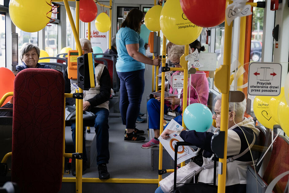 W tramwaju udekorowanym kolorowymi balonami siedzą pasażerowie i czytają ulotki.