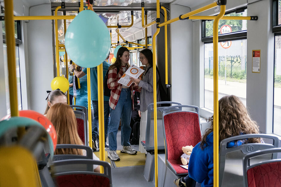 W tramwaju udekorowanym kolorowymi balonami jadą pasażerowie i czytają ulotki.
