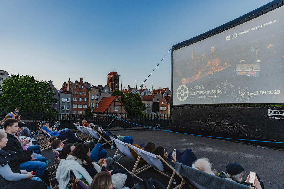 Widok z dachu przedstawia letnie kino plenerowe z dużym ekranem, na którym wyświetlana jest informacja o wydarzeniu Kino na Szekspirowskim  odbywającym się w dniach 28.05–13.09.2025. W tle widać malowniczą panoramę Gdańska z charakterystyczną zabudową i wieżą kościoła Mariackiego. 