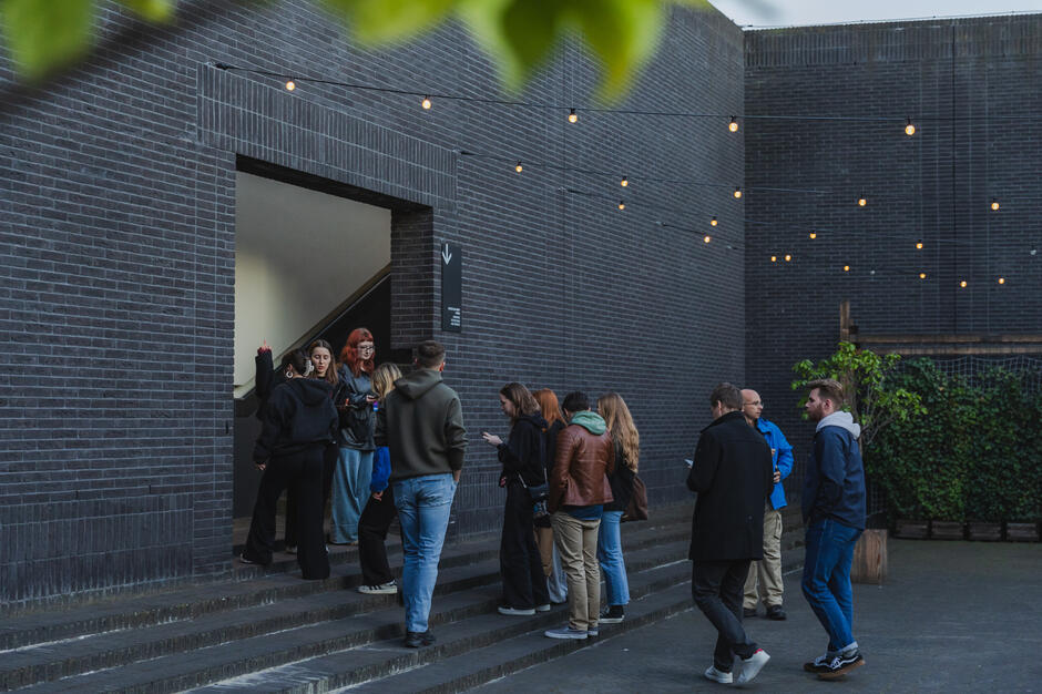 Grupa ludzi czeka w kolejce przed wejściem do budynku z ciemnej cegły, gdzie prawdopodobnie znajduje się wejście na dach Teatru Szekspirowskiego. Nad głowami widoczne są dekoracyjne światełka, które tworzą przytulny klimat wieczornego wydarzenia.

