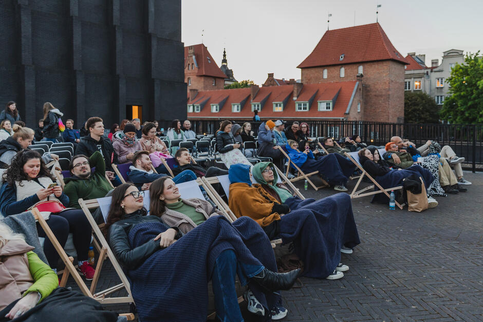 Publiczność zasiadła na leżakach, otulona kocami, gotowa na seans filmowy pod gołym niebem. W tle można dostrzec zabytkową architekturę Gdańska, co dodaje uroku całemu wydarzeniu.