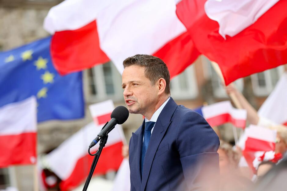 Zdjęcie ponownie przedstawia Rafała Trzaskowskiego przemawiającego na wiecu – w otoczeniu licznych biało-czerwonych flag Polski oraz flag Unii Europejskiej. Jest to dynamiczny i symboliczny kadr, ukazujący go jako lidera przemawiającego do zaangażowanego tłumu.