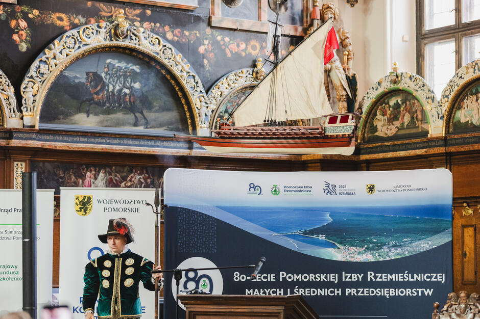 Mężczyzna w stroju halabardnika z epoki stoi koło podwieszonego modelu żaglowca w reprezentacyjnej sali Dworu Artusa.