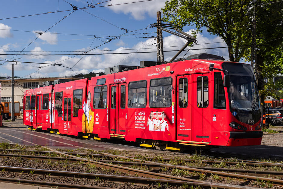 Na zdjęciu widoczny jest czerwony tramwaj należący do Gdańskich Autobusów i Tramwajów, ozdobiony grafiką promującą reprezentację Polski kobiet w piłce nożnej. Tramwaj stoi na torach z siecią trakcyjną nad nim, a w tle widać zabudowania oraz drzewa.