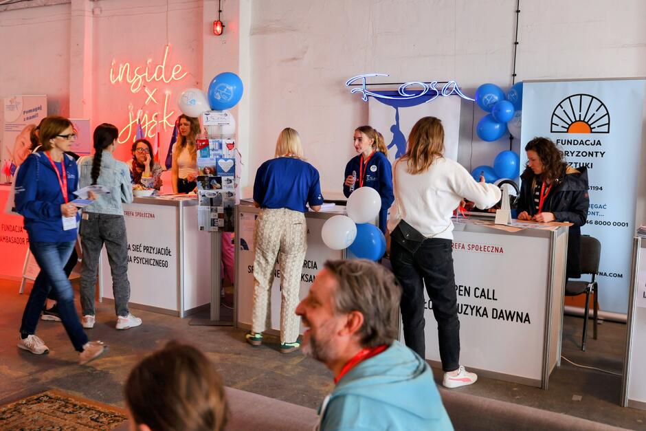 Na zdjęciu widzimy wnętrze strefy informacyjno-promocyjnej, gdzie przedstawiciele różnych fundacji i inicjatyw społecznych rozmawiają z odwiedzającymi przy kolorowych stoiskach. Atmosfera jest żywa i przyjazna, a przestrzeń ozdobiona jest balonami, neonami oraz materiałami promującymi działania na rzecz zdrowia psychicznego i sztuki. 