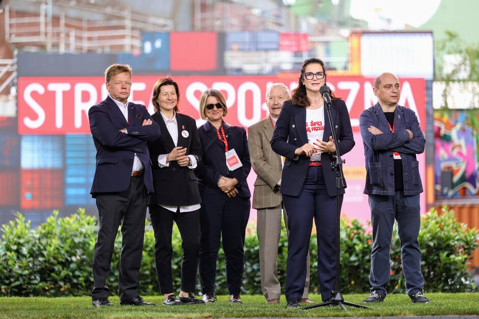 Na zdjęciu widać grupę osób stojących na scenie podczas oficjalnego wydarzenia, a kobieta z mikrofonem na pierwszym planie przemawia do publiczności. W tle znajduje się kolorowa scenografia z napisem „STREFA SPOŁECZNA”, co sugeruje, że wydarzenie ma charakter społeczny lub obywatelski. 