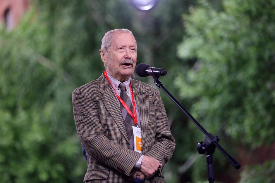 Na zdjęciu starszy mężczyzna w marynarce w kratę przemawia do mikrofonu, stojąc na tle zieleni. Ma przypięty identyfikator i czerwoną smycz, co sugeruje, że bierze udział w oficjalnym wydarzeniu lub konferencji.
