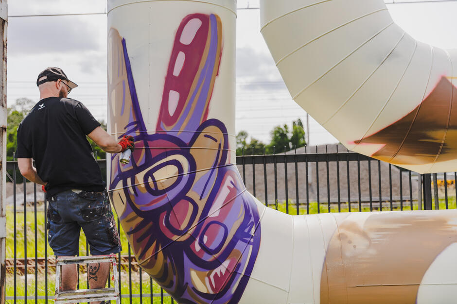 mężczyzna stojący na drabinie i malujący dużą białą rurę z kolorowym projektem graffiti, nosi czarną koszulkę, niebieskie szorty i czapkę baseballową, trzyma pędzel w prawej ręce i wydaje się być w trakcie nakładania graffiti na rurę, w tle znajduje się ogrodzenie i drzewa,
