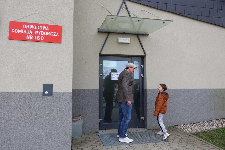 Na zdjęciu widzimy mężczyznę i dziecko stojących przed wejściem do lokalu wyborczego. Na ścianie znajduje się czerwona tablica z napisem: „OBWODOWA KOMISJA WYBORCZA NR 160”