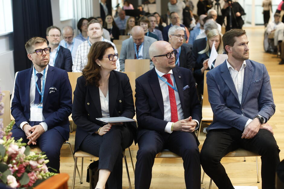 Zdjęcie przedstawia uczestników konferencji siedzących w pierwszym rzędzie, skupionych na prezentacji lub przemówieniu. Osoby są ubrane elegancko, co sugeruje formalny charakter wydarzenia, a sala jest wypełniona słuchaczami.