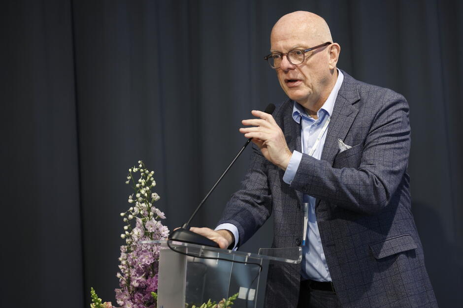 Zdjęcie przedstawia mężczyznę w okularach wygłaszającego przemówienie przy mównicy, ubranego w elegancki, granatowy garnitur. W tle widoczna jest ciemna kotara, a obok mównicy znajduje się dekoracja z kwiatów, co sugeruje oficjalny charakter wydarzenia.