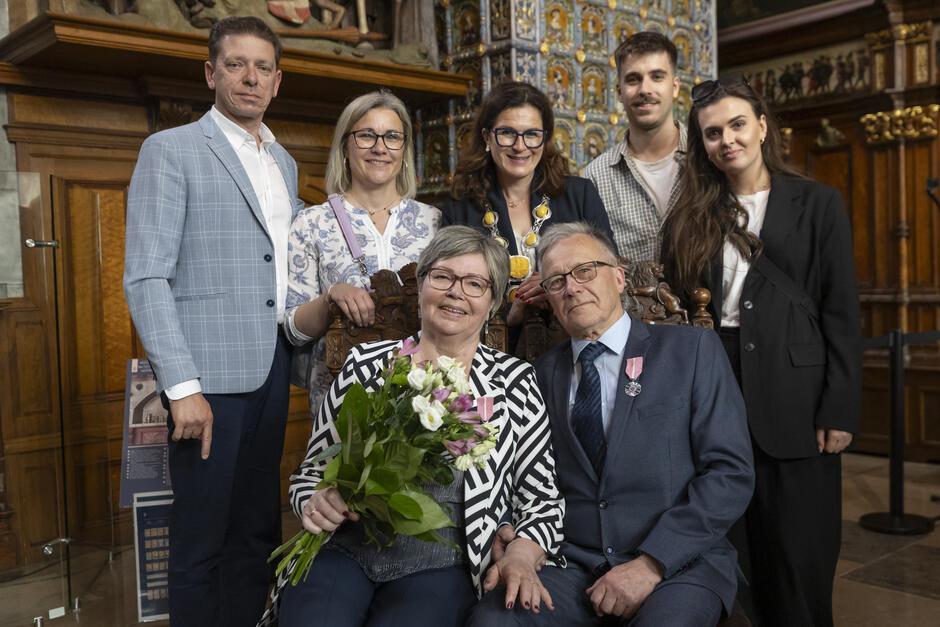 Na tym zdjęciu widzimy starsze małżeństwo siedzące na ozdobnych krzesłach z bukietem kwiatów i medalami, otoczone przez rodzinę w eleganckim wnętrzu. Atmosfera jest uroczysta i pełna dumy, co sugeruje, że para została odznaczona za długoletnie pożycie małżeńskie