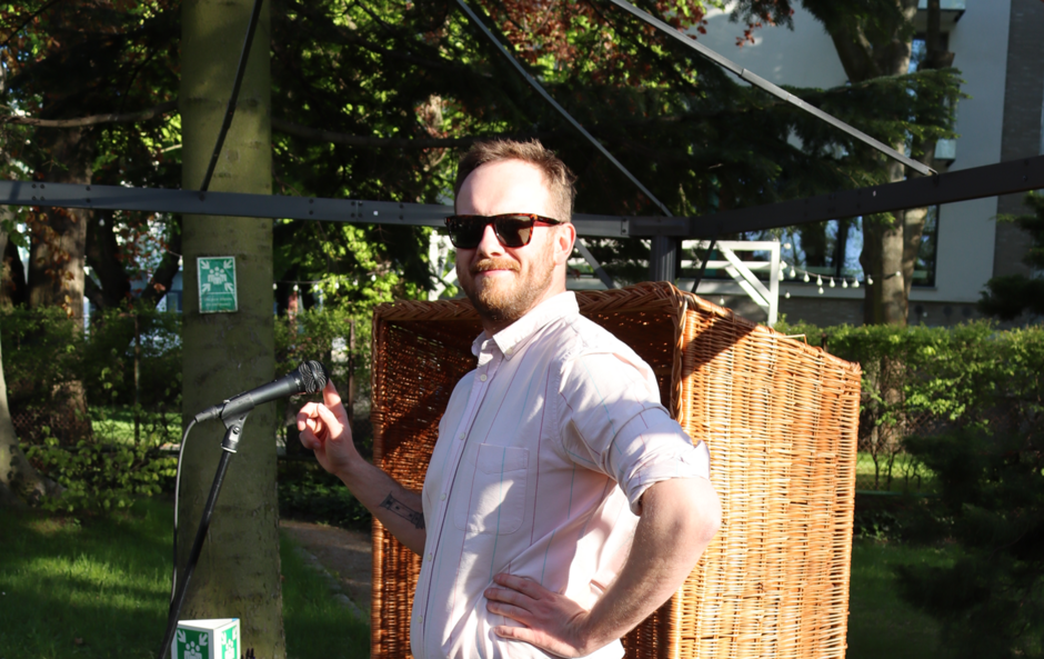 Na zdjęciu mężczyzna z zarostem, w okularach przeciwsłonecznych i koszuli, przed mikrofonem.