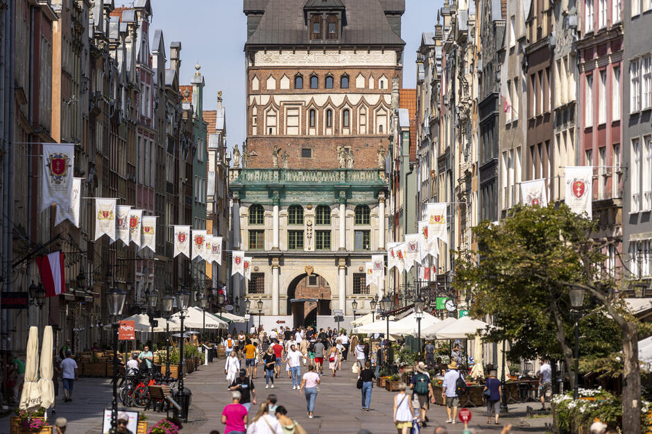 Zdjęcie przedstawia tętniącą życiem ulicę Długą w Gdańsku, pełną spacerujących ludzi oraz kawiarnianych ogródków, ozdobioną flagami z herbem miasta. W tle widoczna jest charakterystyczna Brama Zielona, będąca jednym z najbardziej rozpoznawalnych zabytków miasta.

