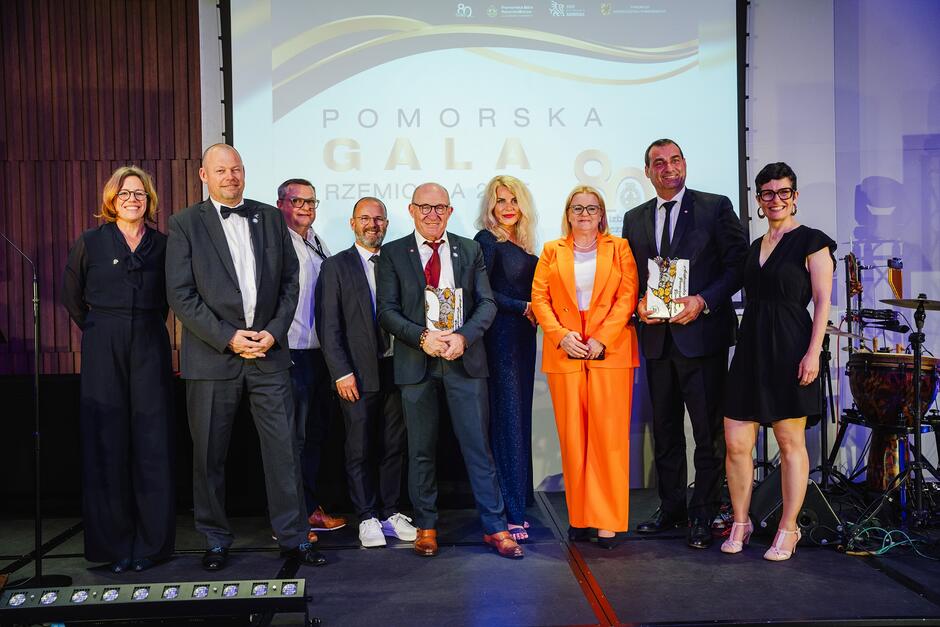 Pięciu mężczyzn i cztery kobiety, wszyscy elegancko ubrani, pozują do fotografii na scenie. Dwóch mężczyzn trzyma w ręku statuetki - nagrody. Za nimi ekran z napisem Pomorska Gala Rzemiosła .