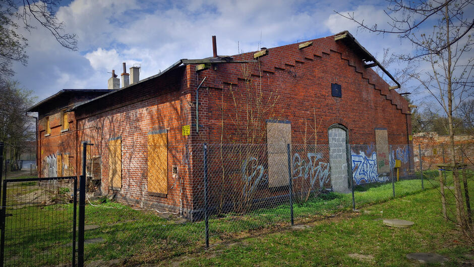 Na zdjęciu widoczny jest stary, ceglany budynek o przemysłowym charakterze, otoczony ogrodzeniem z siatki i porośnięty częściowo graffiti. Obiekt jest opuszczony, z zabitymi deskami oknami i zamurowanym wejściem, co świadczy o jego długoletnim braku użytkowania.