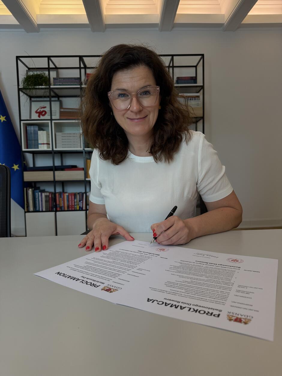 Na zdjęciu kobieta w białej bluzce, siedząca przy biurku, podpisuje dokument zatytułowany „Proklamacja Światowego Dnia Roweru” z herbem Gdańska. W tle widać półkę z książkami i flagę Unii Europejskiej, co sugeruje oficjalny charakter miejsca i wydarzenia.