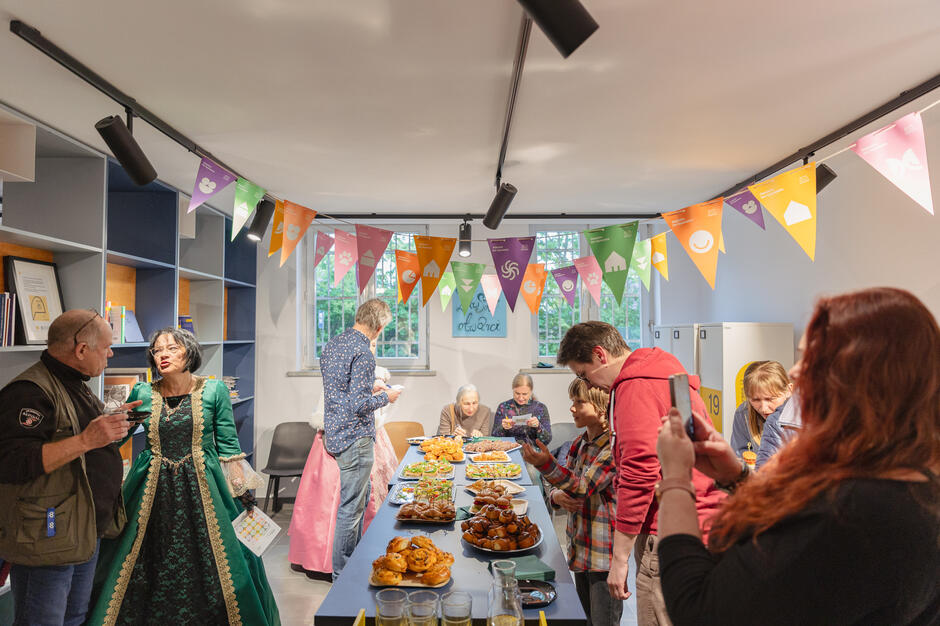 W sali stoją wokół stołu ludzie, niektórzy przebrani w stroje z epoki, i rozmawiają. na stołach jedzenie. Pod sufitem wiszą proporczyki. 