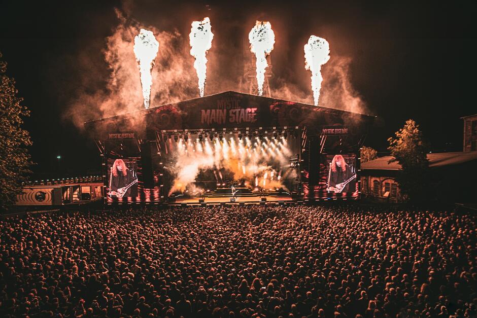 Zdjęcie przedstawia koncert na dużej scenie głównej festiwalu Mystic Festival, z efektownymi wybuchami ognia, intensywnym oświetleniem i ogromnym tłumem fanów przed sceną. Na dwóch bocznych ekranach widać gitarzystę w trakcie występu, co potęguje atmosferę energetycznego widowiska