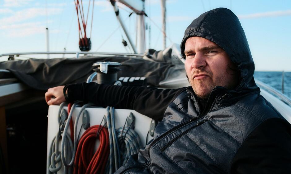 Na zdjęciu mężczyzna w kapturze, znajduje się na pokładzie statku, w tle morze. 