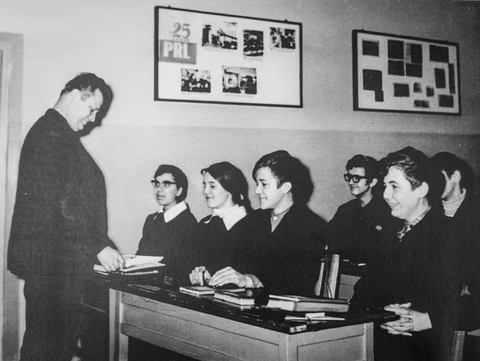 To czarno-białe zdjęcie przedstawia scenę z lekcji szkolnej w czasach PRL-u – prawdopodobnie lata 70. lub wczesne 80. XX wieku. Fotografia została wykonana w klasie, w której uczniowie siedzą w drewnianych ławkach, a nauczyciel rozmawia z uczennicą przy pierwszej ławce. W tle widoczna jest dekoracja ścienna z napisem „25 lat PRL”, co wskazuje na rocznicę obchodzoną w 1969 roku