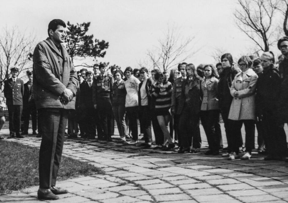 To czarno-białe zdjęcie przedstawia nauczyciela lub przewodnika przemawiającego do grupy młodzieży, najprawdopodobniej uczniów III Liceum Ogólnokształcącego w Gdańsku. Zdjęcie wykonano na zewnątrz – być może podczas szkolnej wycieczki, rajdu lub uroczystości patriotycznej.