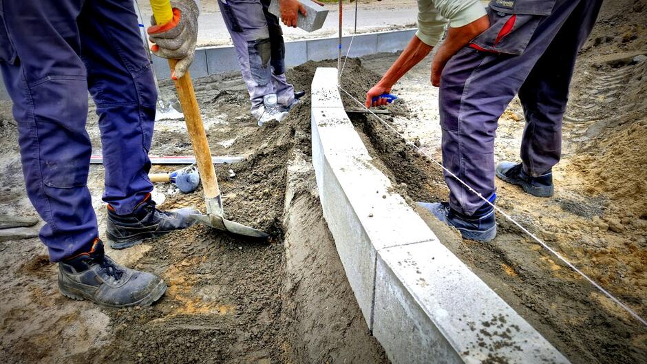 na zdjęciu dwóch robotników w roboczych strojach układa krawężnik na gruntowej drodze